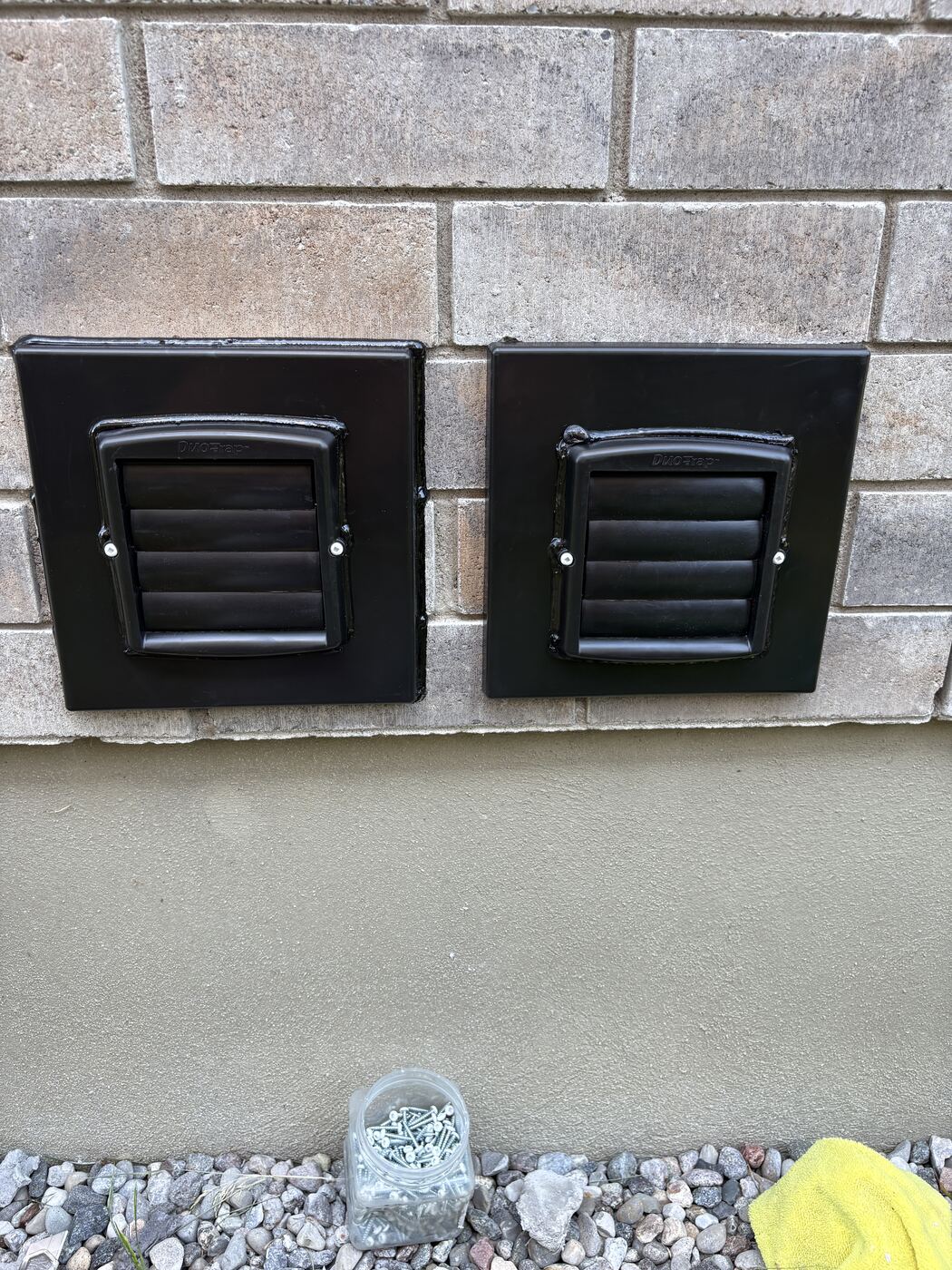Two black louvered Thermo-Trap vents mounted on black plates over brick