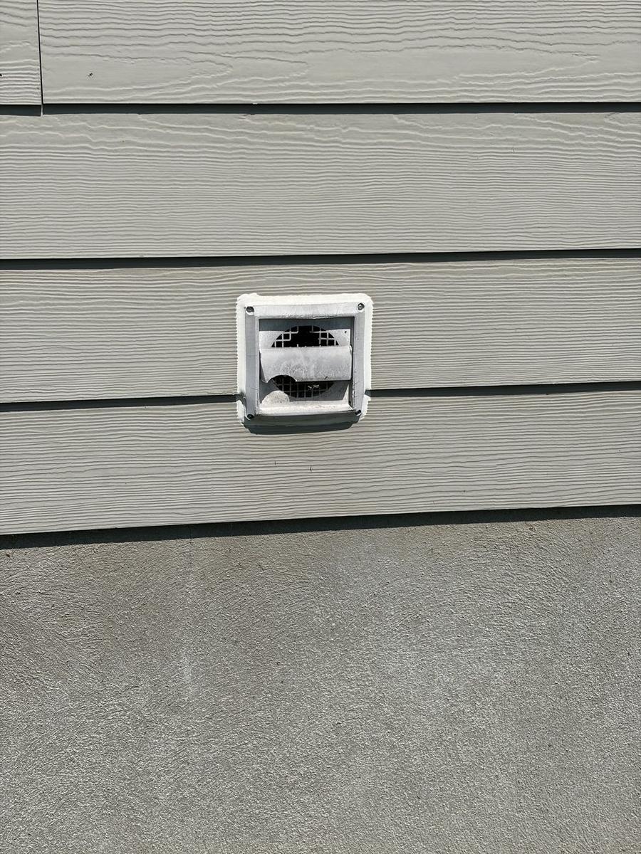 Cracked exterior dryer vent cover with a broken flap hanging open on light siding