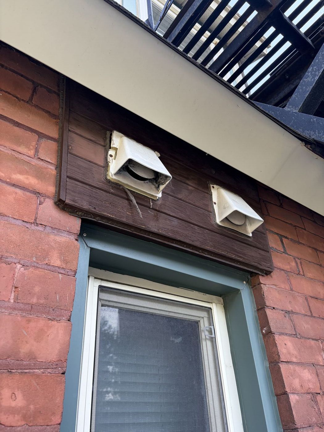 Two old exterior vent covers on a wood panel above a window on a brick wall